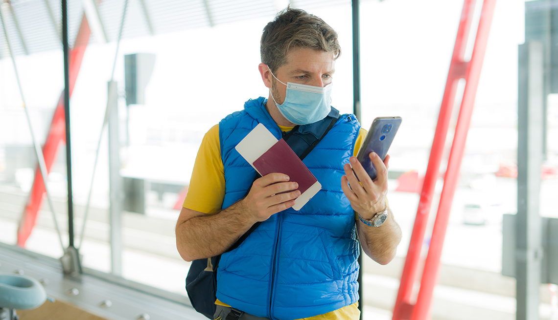 Un hombre con una máscara y un chaleco azul en el aeropuerto mira su teléfono
