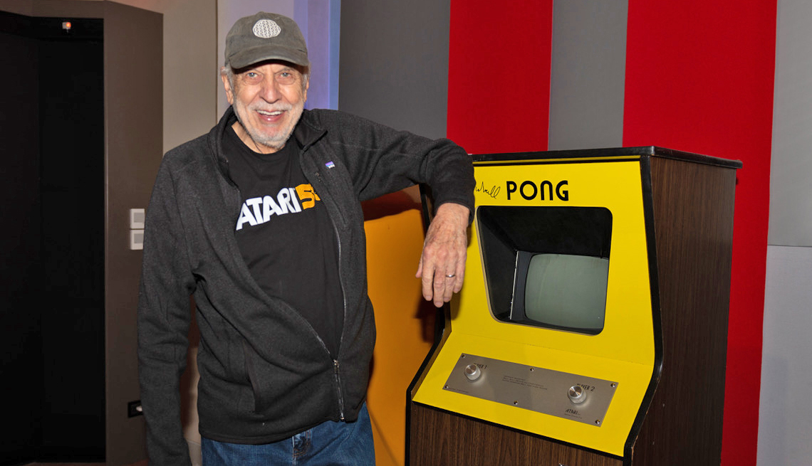 atari co-founder nolan bushnell wears a company t-shirt and a black jacket while leaning on an arcade-style pong machine