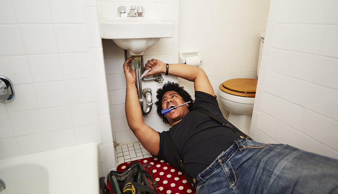 A man is laying on a bathroom floor, looking up at a sink and holding a tool around a pipe