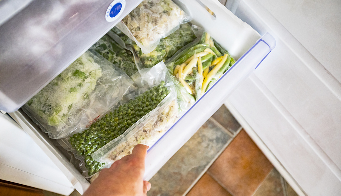 Una mano abre el cajón del congelador de una nevera lleno de vegetales