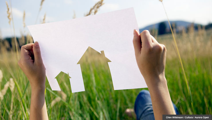 A person holding up a paper cut-out of a house in front of a grassy field.