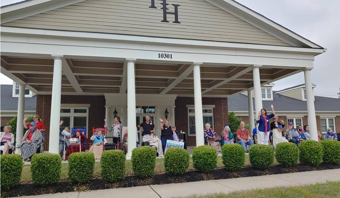 Nursing home residents gather beneath a covered porch to watch a parade