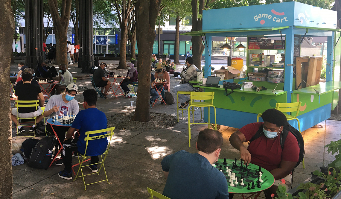 People play chess on outdoor tables in a park with a Game Cart kiosk
