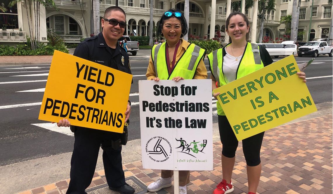 A police officer and two women hold signs promoting pedestrian safety