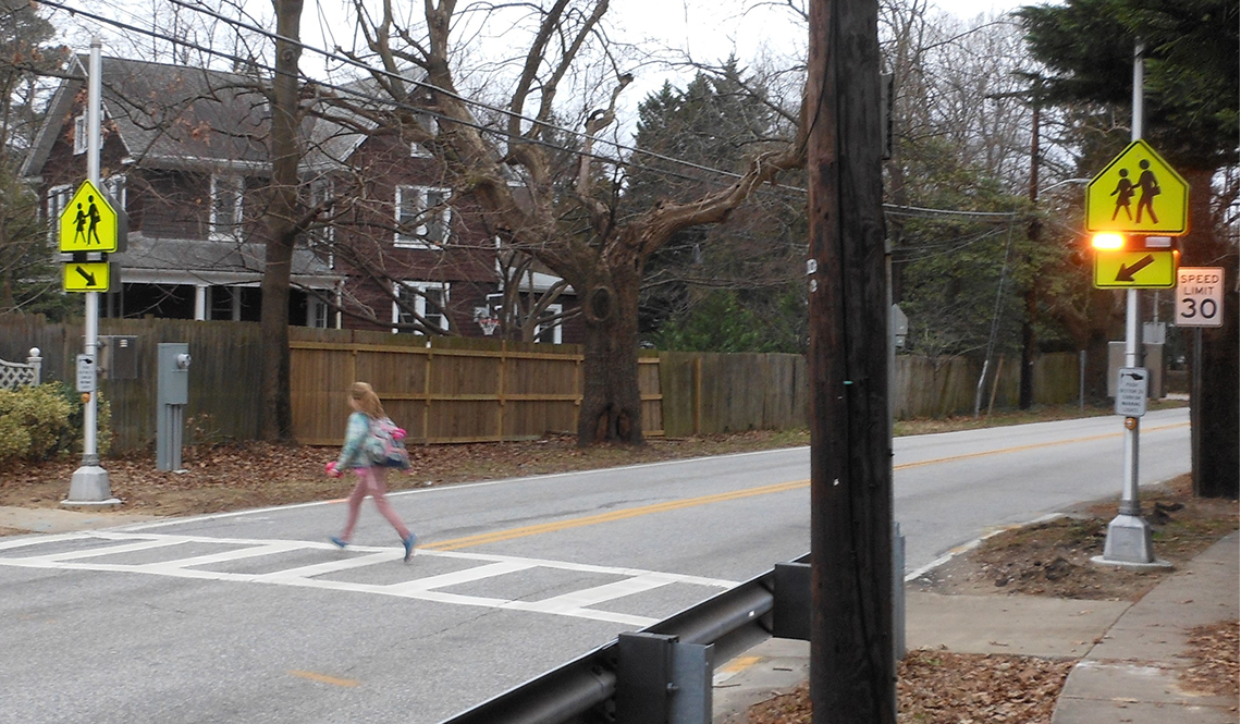 A lighted pedestrian beacon, school crossing signage and a crosswalk help a young girl safely cross a tow-lane street