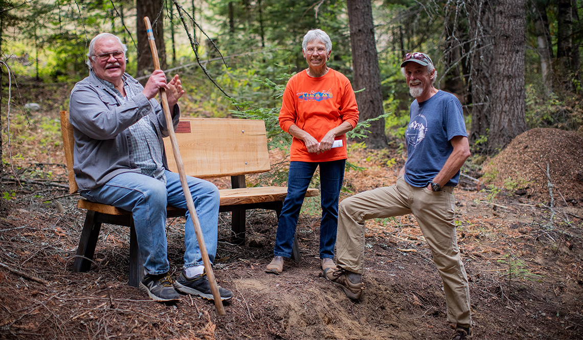 Three older adults pose with a new wooden bench installed in an Idaho forest