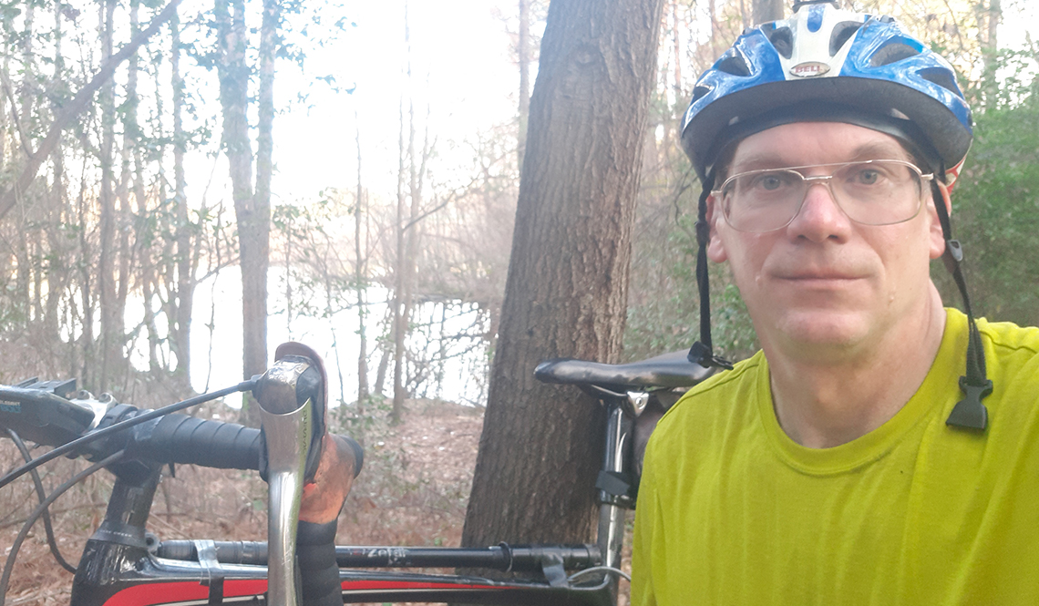 David Maryniak taking a break from a bike ride in a woods near a lake