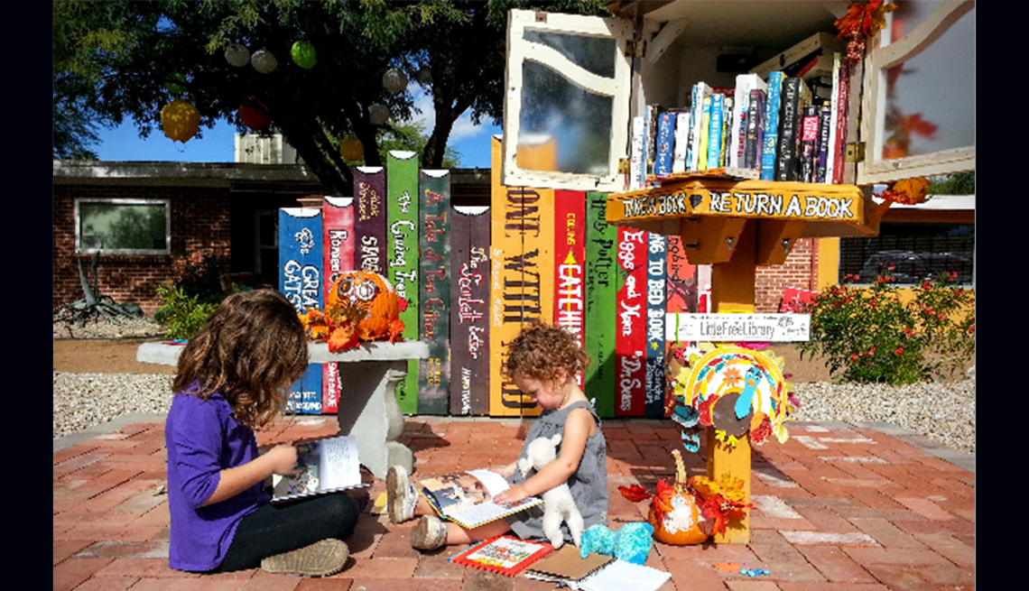 A young girl and a girl toddler sit on a brick path and read books in front of a little library.