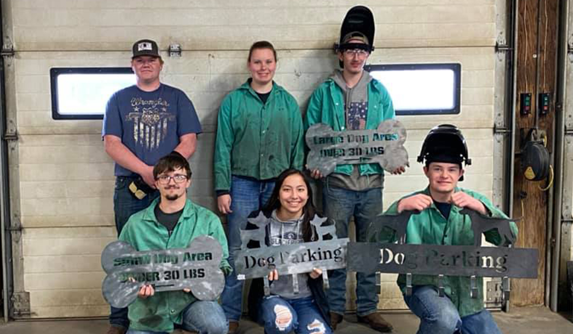 Six people pose in a metal shop with metal signs made for the City Bark Park