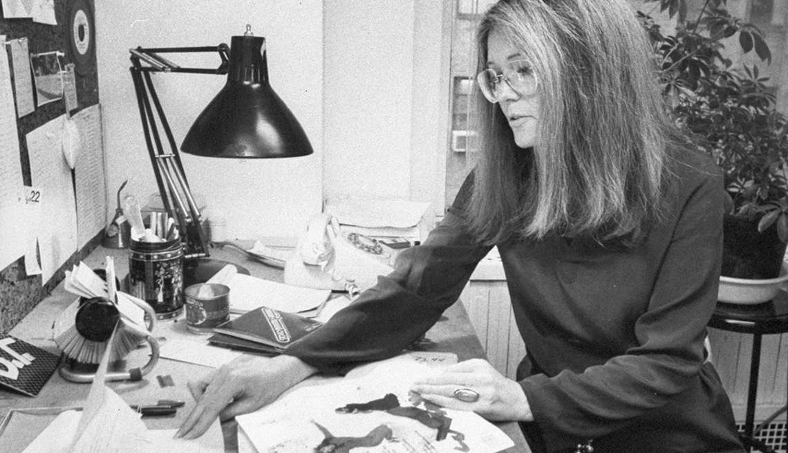 item 12 of Gallery image - Gloria Steinem sits at a desk with a lamp on it reading