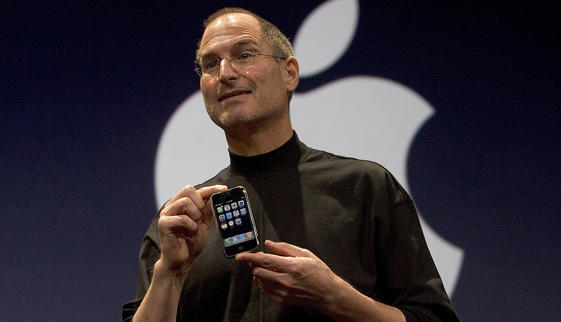 item 23 of Gallery image - The late CEO of Apple, Steve Jobs, is wearing black turtleneck and holding an iPhone between his forefingers in front of the company logo