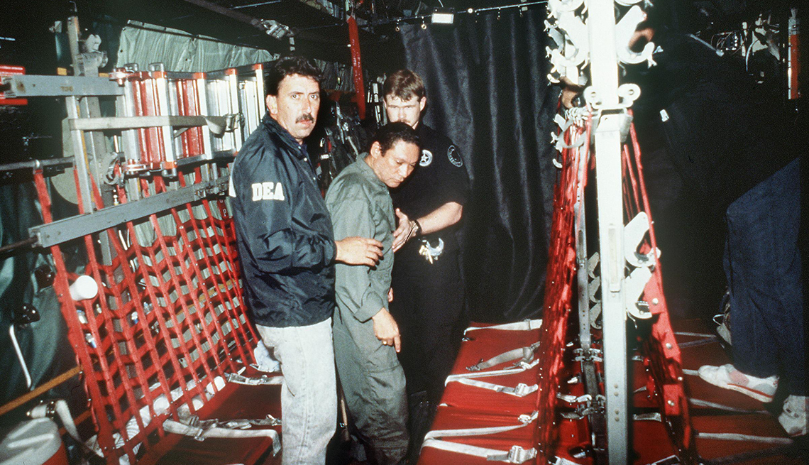 item 29 of Gallery image - Two men in uniforms hold onto Manuel Noriega near red seats on an aircraft