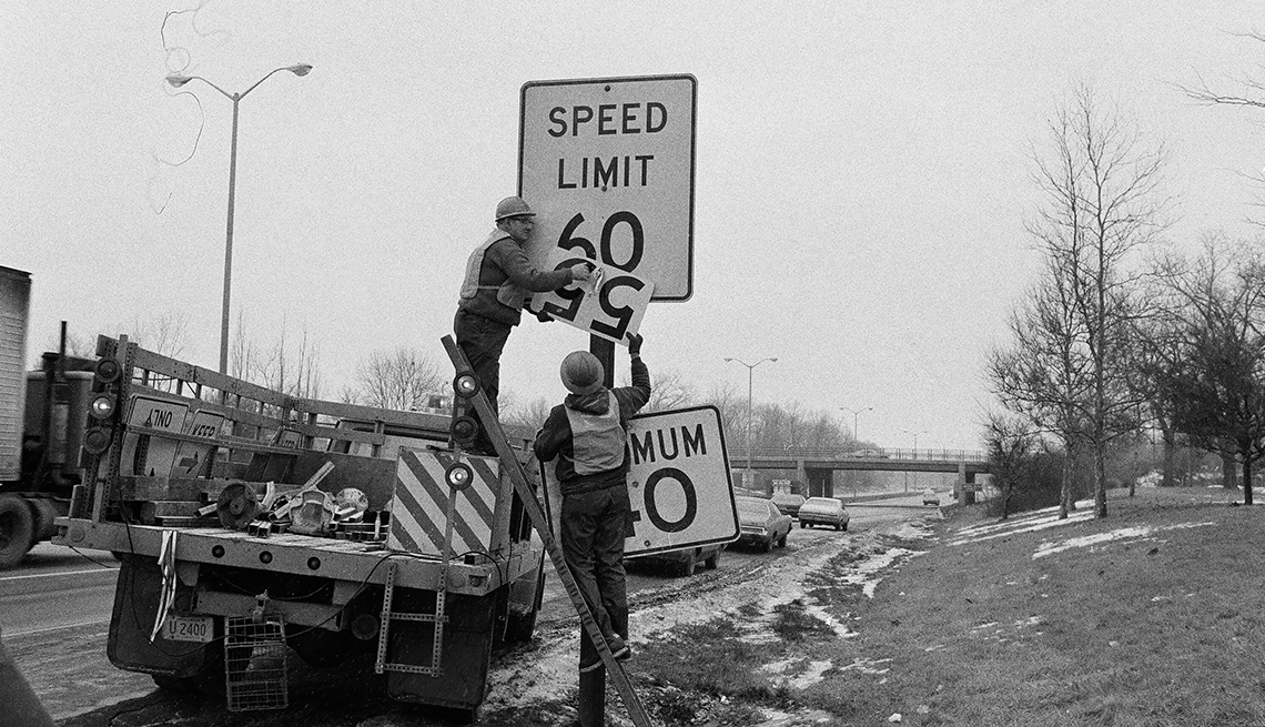 item 30 of Gallery image - One worker stands in a truck bed while another stands to his right as they replace a speed limit sign that says 50 with another that says 55