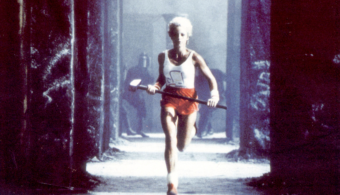 item 10 of Gallery image - A blonde woman in a white tank top and red short carries a sledgehammer while she runs between sets of black columns with black-clad police chasing her in Apple's 1984 commercial