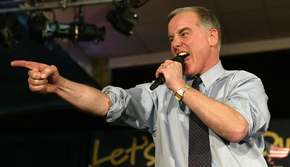 Howard Dean is wearing a tie with his shirtsleeves rolled up while he points and shouts into a microphone