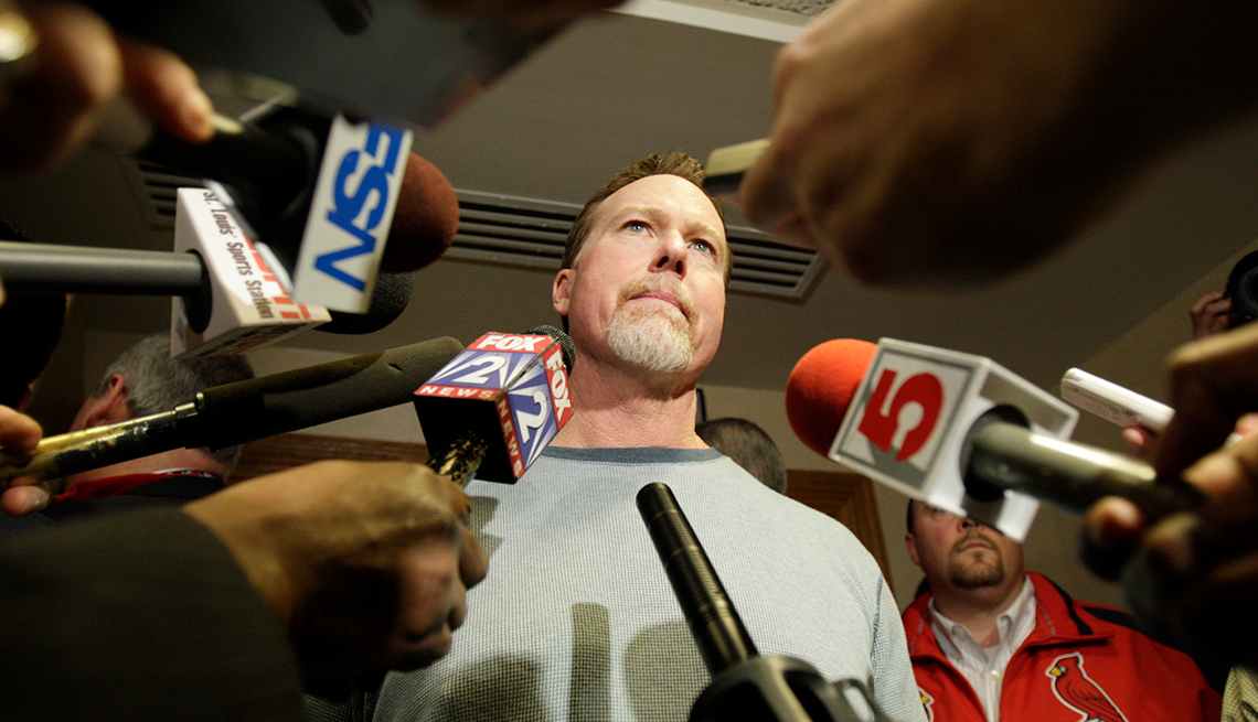 St. Louis Cardinals baseball player Mark McGwire frowns while being surrounded by microphones as a team staff member looks on behind him