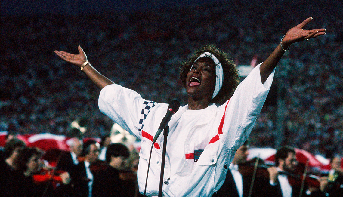 Whitney Houston in a white headband and a white and red windbreaker raises her arms while singing the National Anthem at the Super Bowl