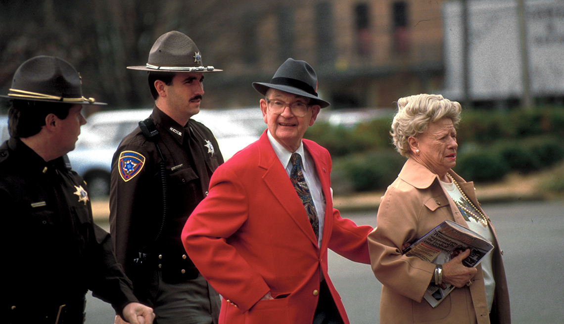 item 24 of Gallery image - Two state troopers in brown uniforms follow Byron De La Beckwith who's wearing a red jacket, a tie and a fedora, while taking the arm of a woman in a beige coat holding a bundle of papers