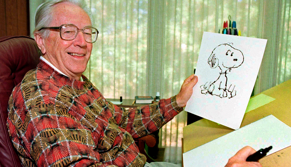 item 16 of Gallery image - cartoonist Charles M. Schultz is smiling while holding a drawing of Snoopy in one hand and a pen in the other