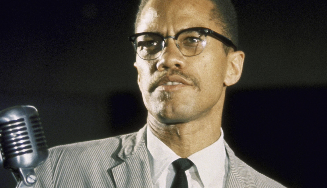 item 8 of Gallery image - malcom x in a gray suit and black tie behind a microphone