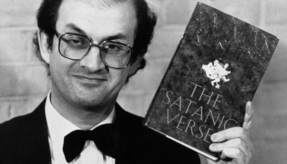 item 15 of Gallery image - a black and white picture of author Salman Rushdie holding up a copy of his book, The Satanic Verses