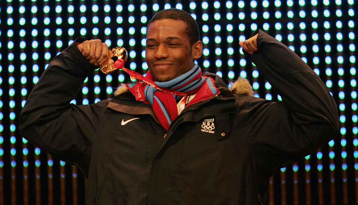 item 11 of Gallery image - Olympian Shani Davis is smiling and showing off a gold medal while wearing a blue and red scarf and a black Team USA winter jacket in front of a black background studded with lights