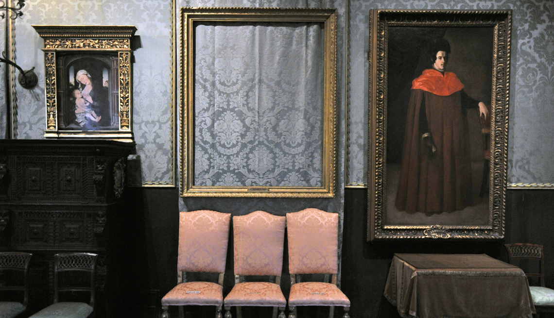 item 14 of Gallery image - an empty frame and two paintings hang on a gray cloth-covered wall behind three chairs