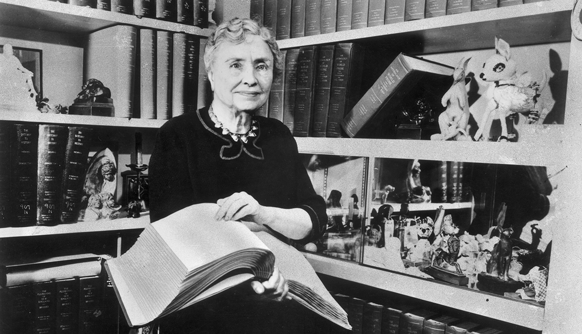 portrait of helen keller holding a braille volume and surrounded by shelves containing books and decorative figurines