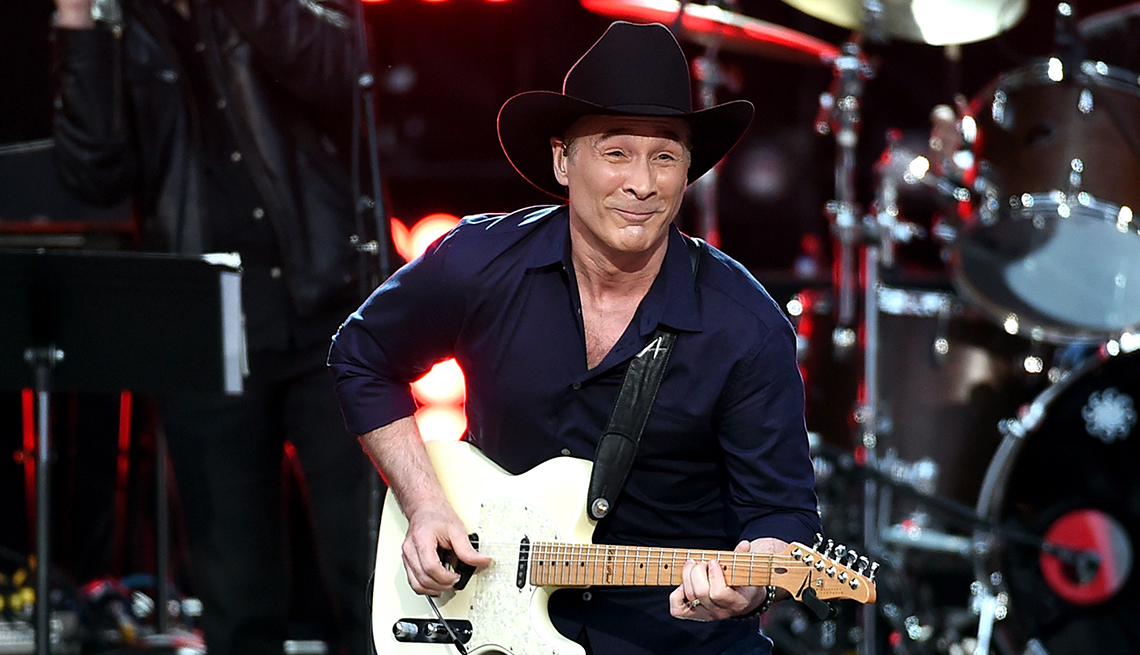 Clint Black onstage playing white electric guitar with drum set and another musician behind him