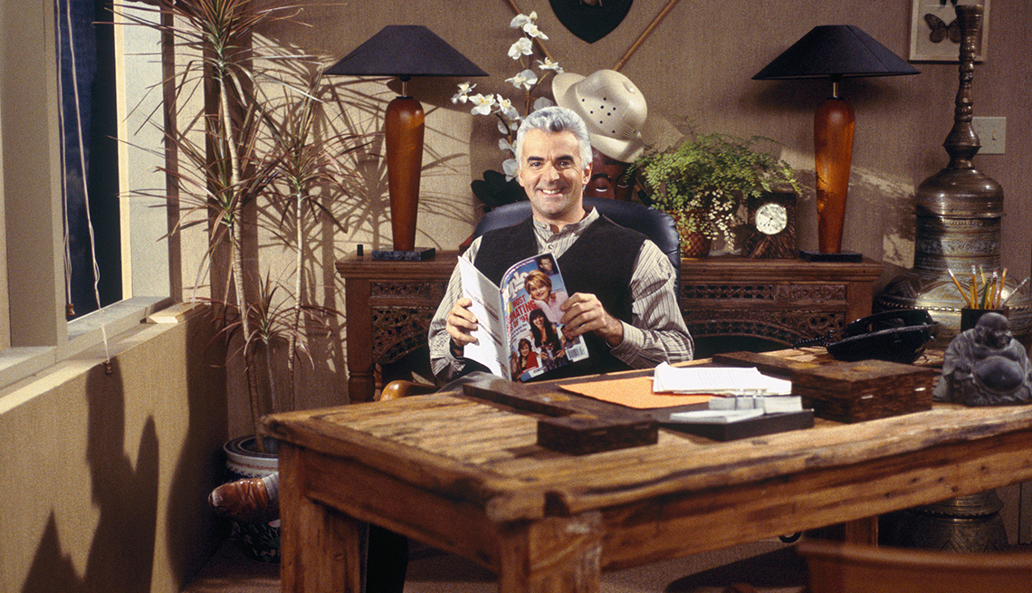 John O'Hurley as J. Peterman on a 'Seinfeld' episode, sitting at a desk smiling and holding a magazine