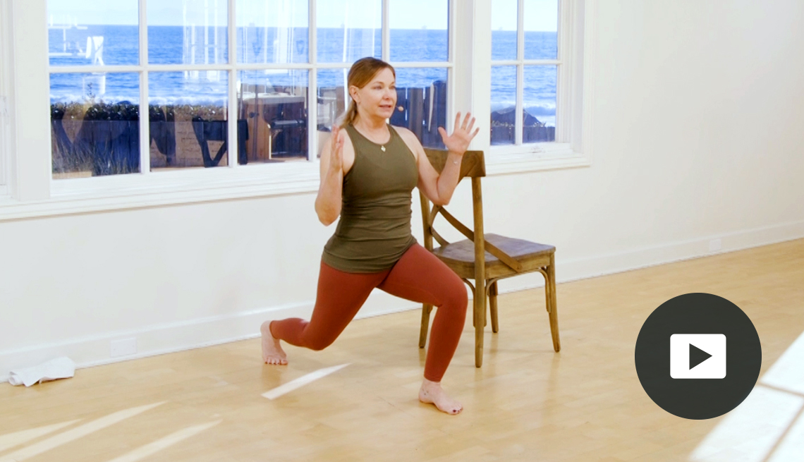 Amy Havens doing pilates in a studio with a big window, standing beside a chair in a lunge position, with video icon overlay