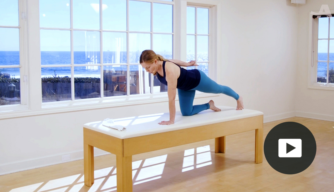 woman in Pilates pose in a studio on a bench in front of a window, and video icon overlay