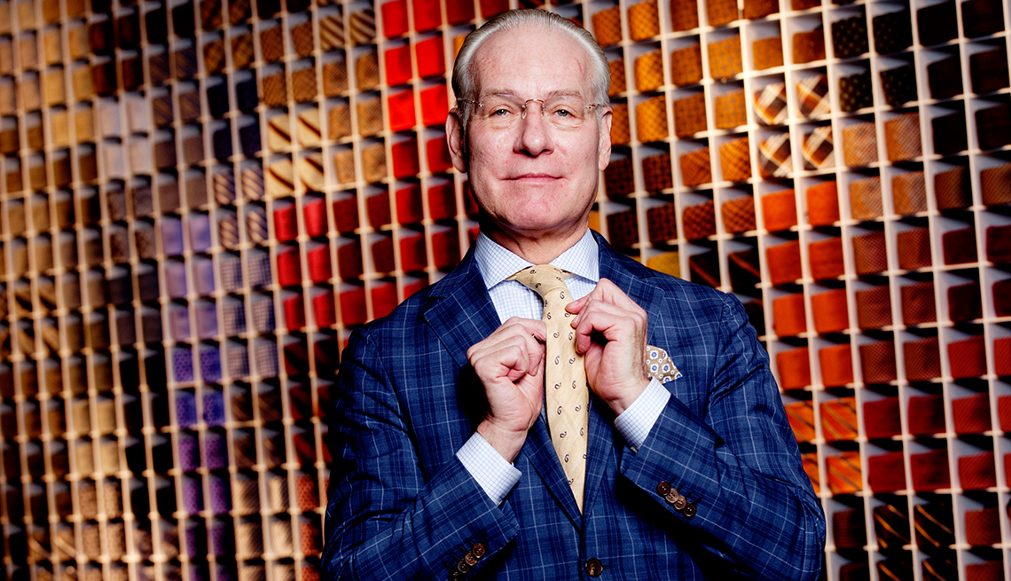 Tim Gunn holding his tie while standing in front of a huge wall display of multicolored ties