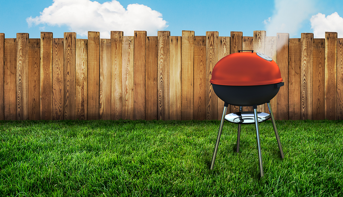 a grill with smoke emanating from its red cover outside on green grass in front of a wood fence and blue skies