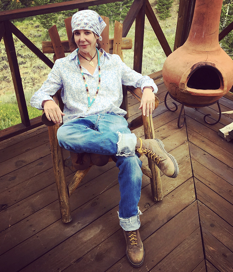 author Tracy Achor Hayes wearing hiking boots and casual cloths, sitting on deck of cabin next to wood-burning stove 