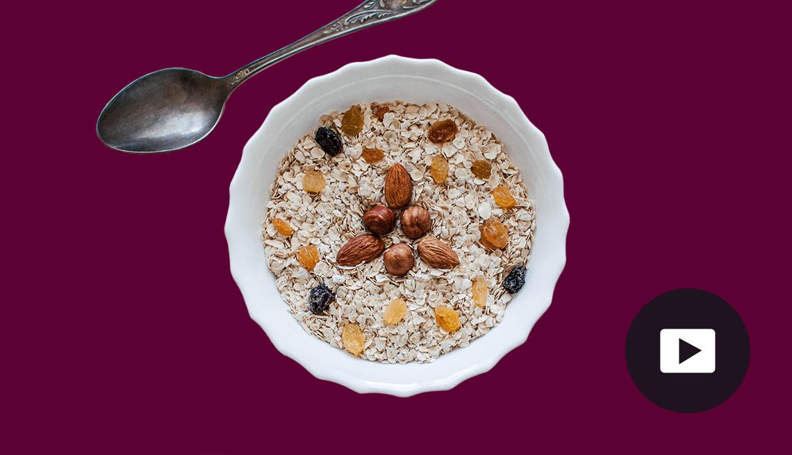 A bowl of oat cereal decorated with nuts and raisins. Metal spoon sits beside the bowl. Dark background. Video play button in lower right corner.
