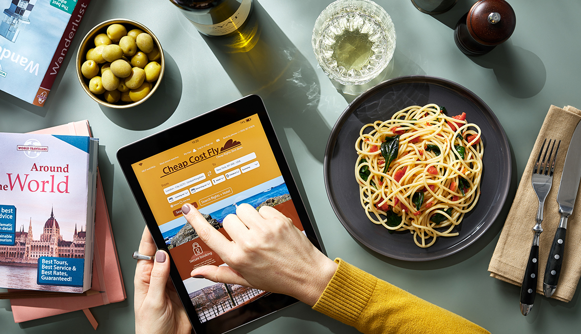 bowl of olives, woman's hands operating a tablet with "Cheap Cost Fly" webpage on screen, plate of pasta, fork, knife, travel books, wine bottle, crystal drinking glass