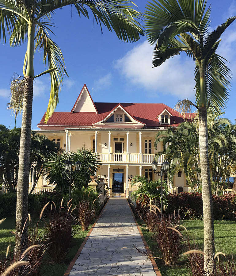 A front view of Domaine Saint-Aubin, a former plantation house turned hotel on the coast of Martinique. The house is surrounded by palm trees. 