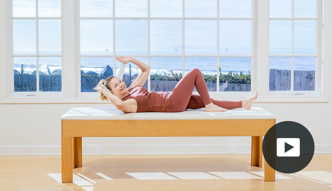 Pilates instructor Amy Havens uses an elastic band to exercise on a padded table in front of a window with a view of a fence, rocks and the ocean. Video play button in lower right corner 