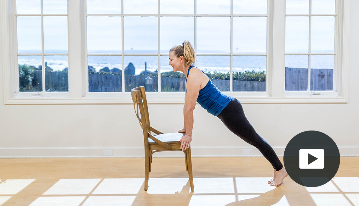 Pilates instructor Amy Havens exercises with a wooden chair in front of a window with a view of rocks, a fence and the ocean. Video play button in lower right corner.