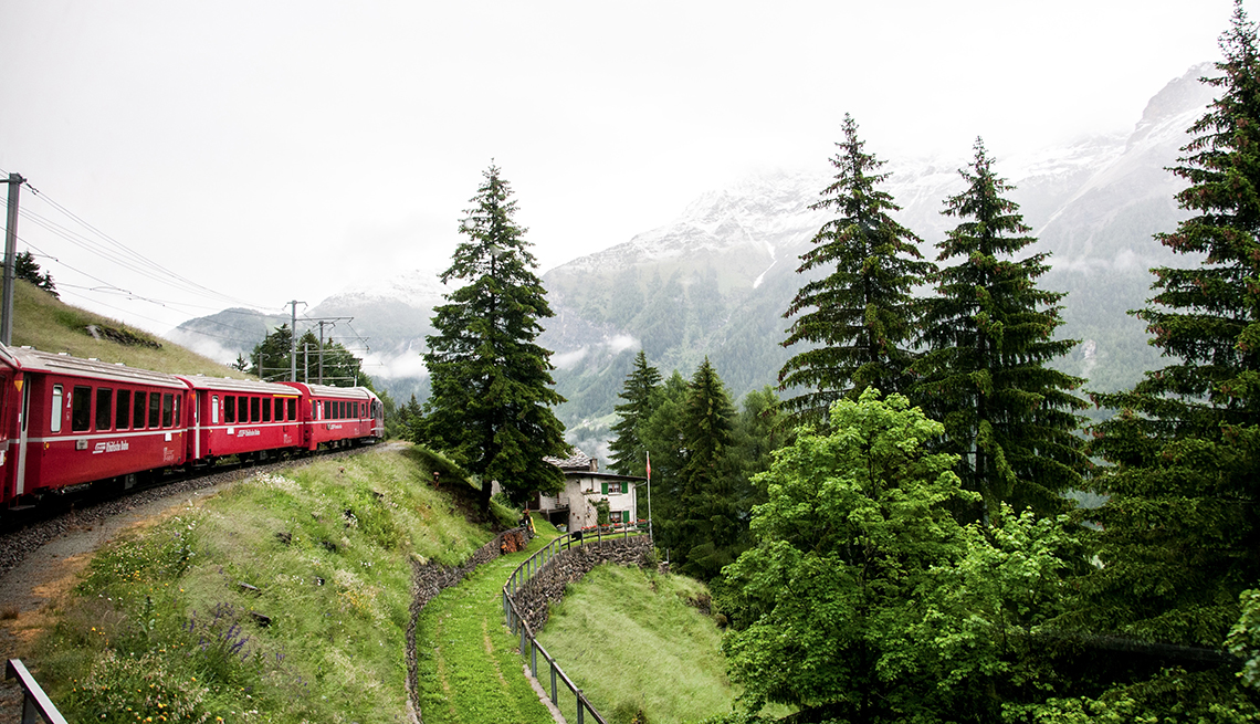 A Bernina Red Train travels through the Swiss Alps. Trees and snow-capped mountains are along the route
