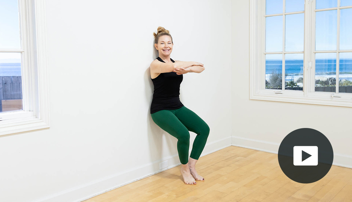 Pilates instructor Amy Havens squats against a wall while barefoot on a hardwood floor. A window to the side shows trees and a body of water outside the room. Video play button in lower right corner.