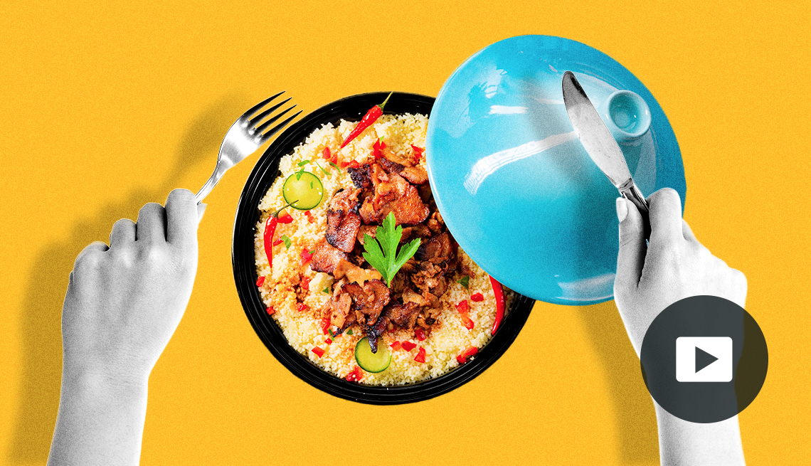 A black bowl of Moroccan chicken with a small plate. A person’s hands hold a fork and knife. Yellow background. Video play button in lower right corner.