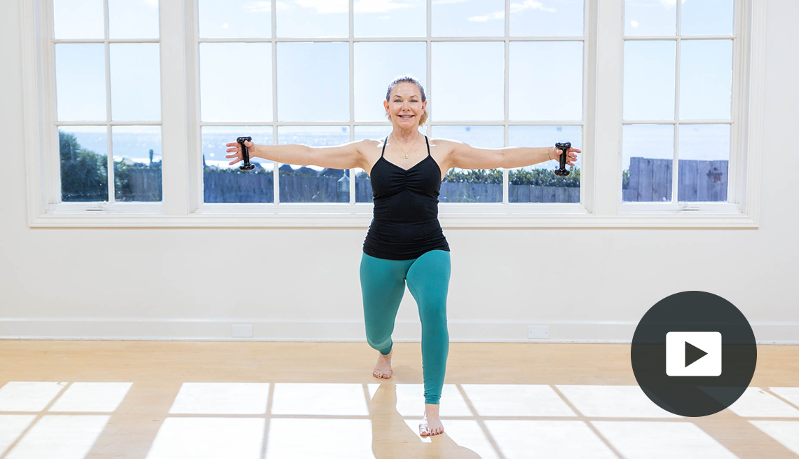 Pilates instructor Amy Havens holds small dumbbells out to her sides as her lunges forward. She is standing in front of windows that show an outdoor water scene. Video player icon in lower right corner.