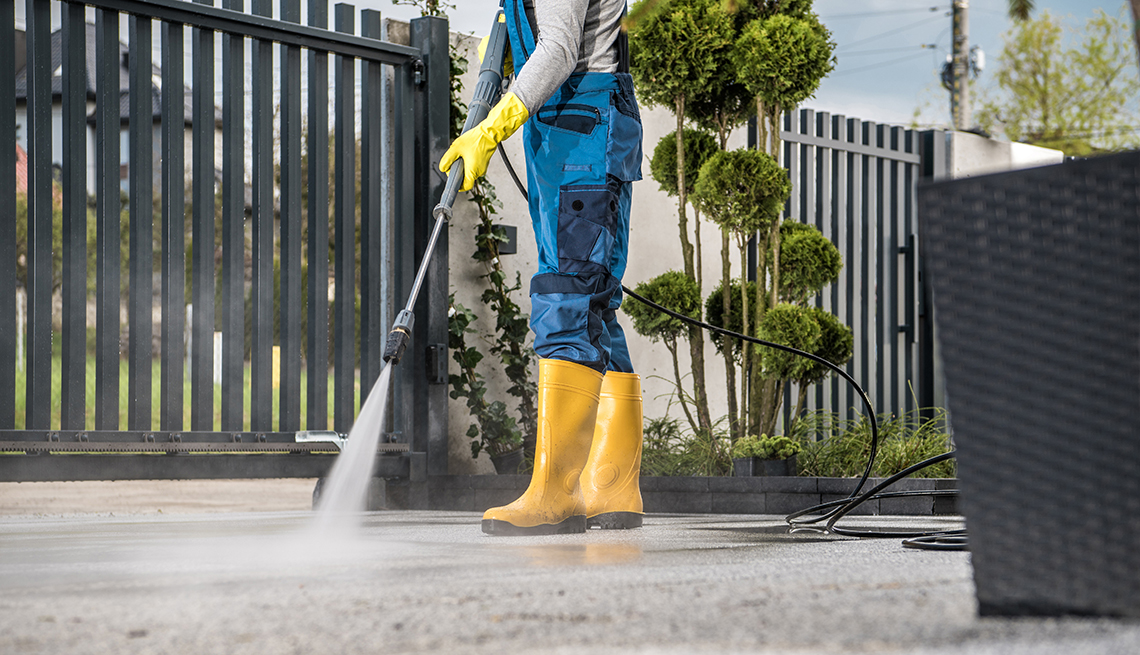 person wearing yellow rain boots and work uniform pressure washing concrete next to black gate and small tree