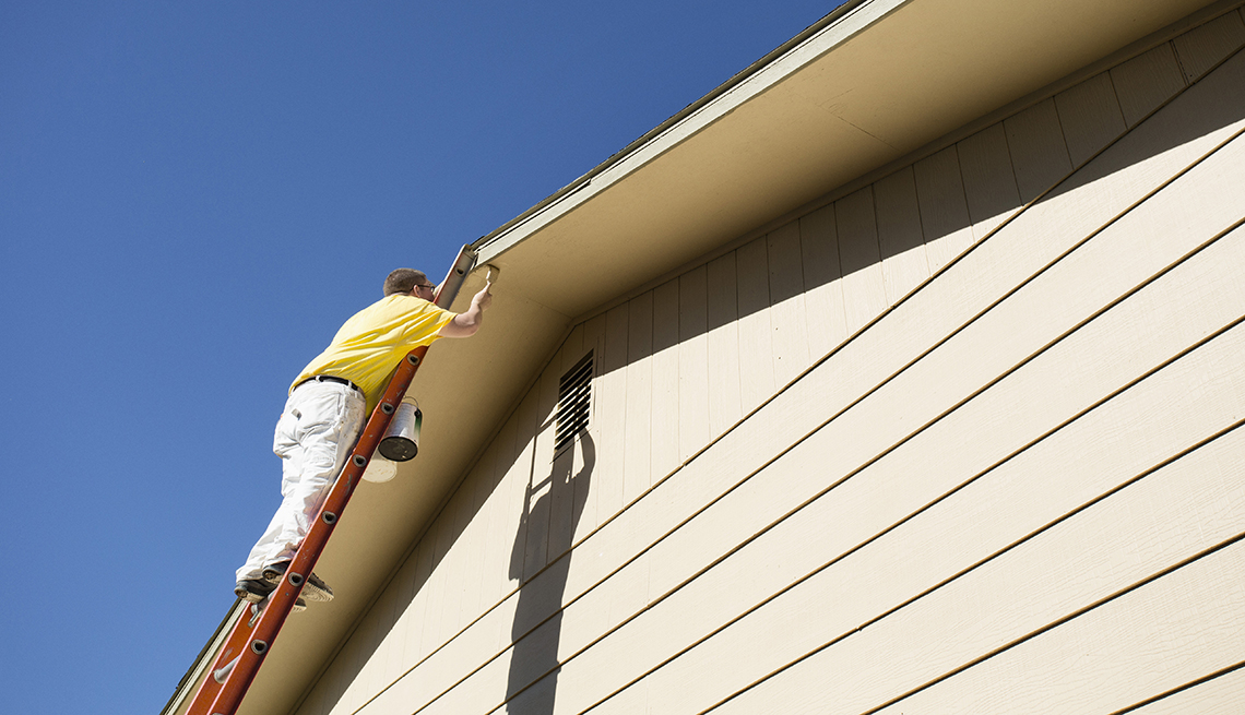 man on orange ladder painting edge of roof of tan house