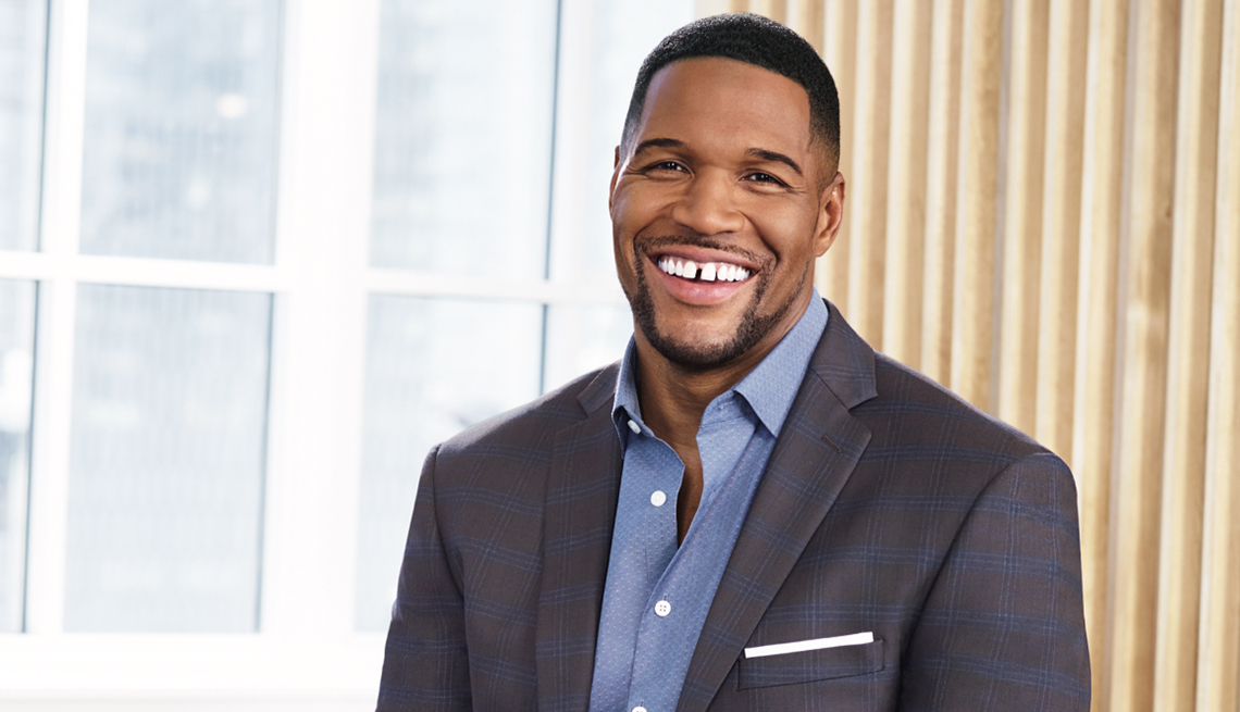 michael strahan smiling, wearing a blue button down shirt and grey suit jacket in front of window with white trim