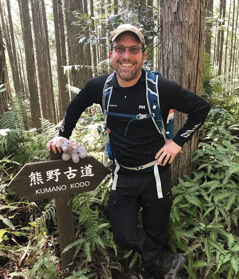 david hochman holding small stuffed elephant, standing next to sign that reads kumano kodo in the woods, surrounded by trees
