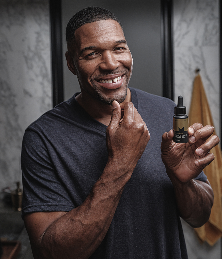 michael strahan touching chin and holding bottle of beard oil from his skin care line with the other hand, towel hanging up against marble wall behind him