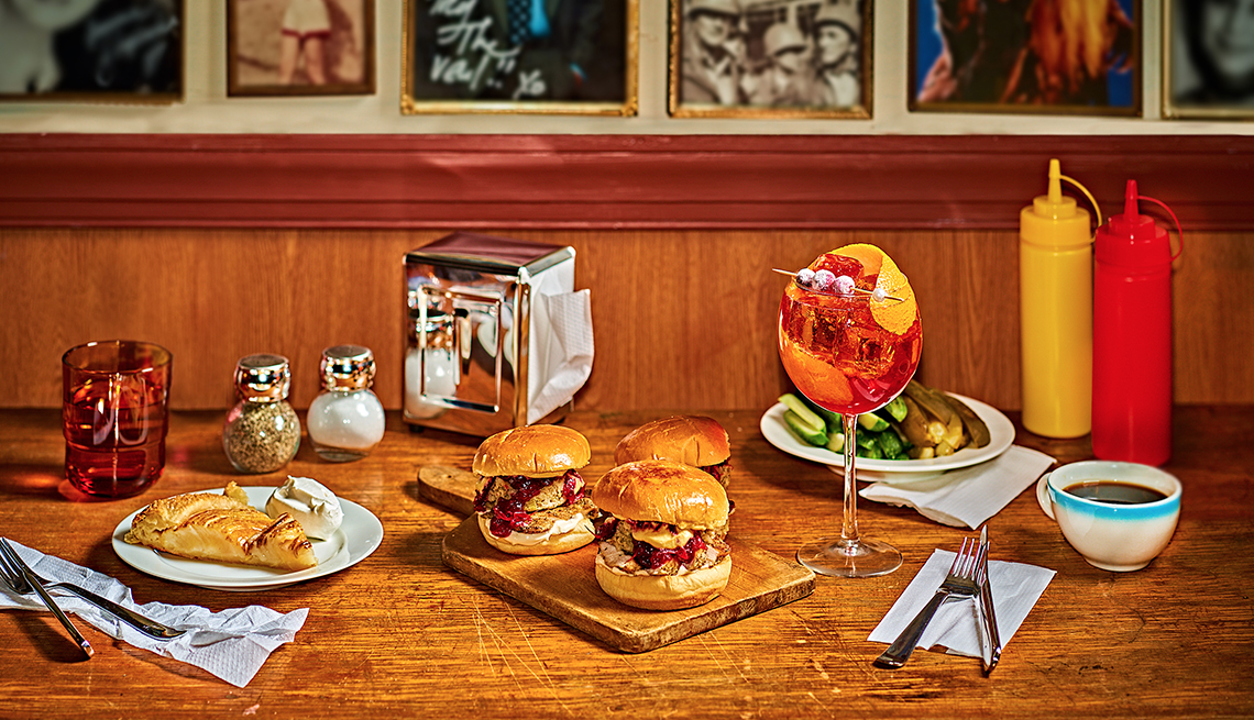 wood table set with three gobbler sandwiches, a slice of pear galette, stem glass of cranberry aperol spritz, a cup of coffee, plate of pickles, glass of water, mustard and ketchup, salt and pepper, restaurant napkin dispenser, and a row of framed pictures in the background typical of diner decor